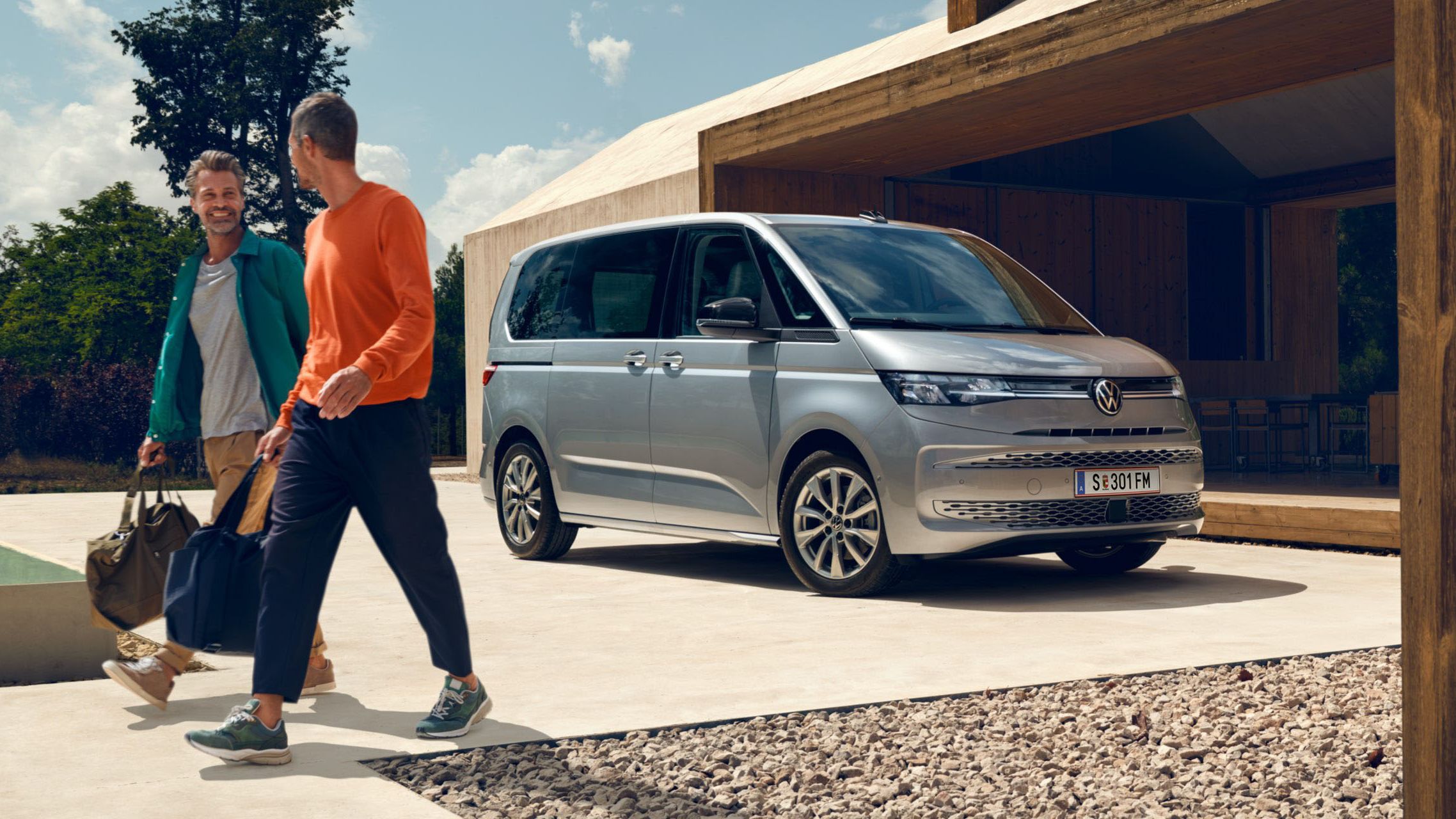 Zwei Männer mit Sporttaschen laufen vor einem neuen silbernen VW Multivan T7 2021.